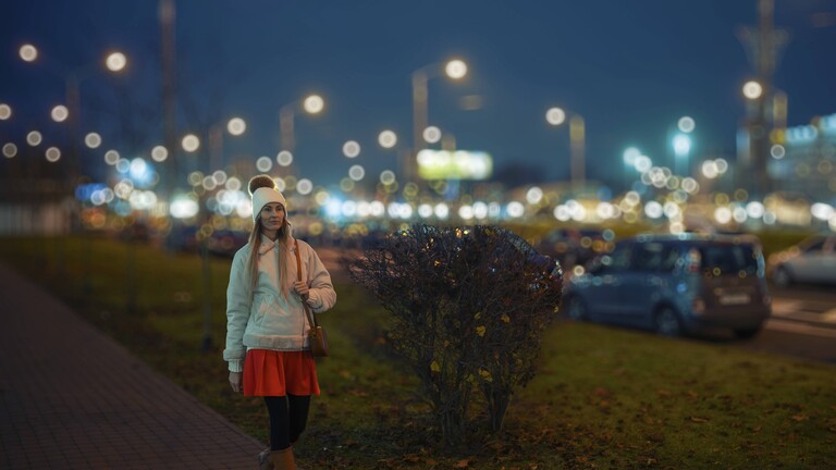 Eine Frau geht nachts eine Stadtstraße entlang und trägt ein Outfit, das einen kurzen Mantel, eine Strickmütze und einen kurzen Rock umfasst. Straßenlaternen und festliche Lichter schaffen eine lebendige Atmosphäre um sie herum.
