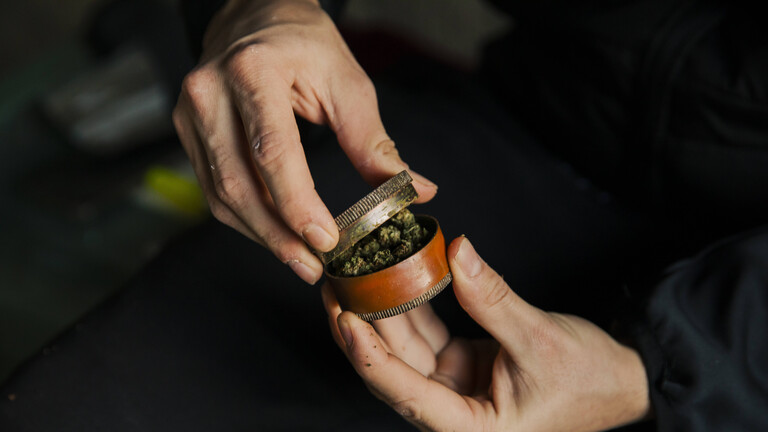 Zwei Hände halten eine kleine Dose in der Hand, der Deckel ist leicht geöffnet. In der Dose ist Marihuana.