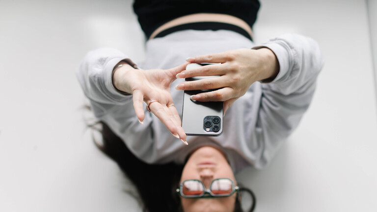 Eine junge Frau liegt auf dem Rücken und tippt auf ihrem Smartphone, das sie über sich hält.
