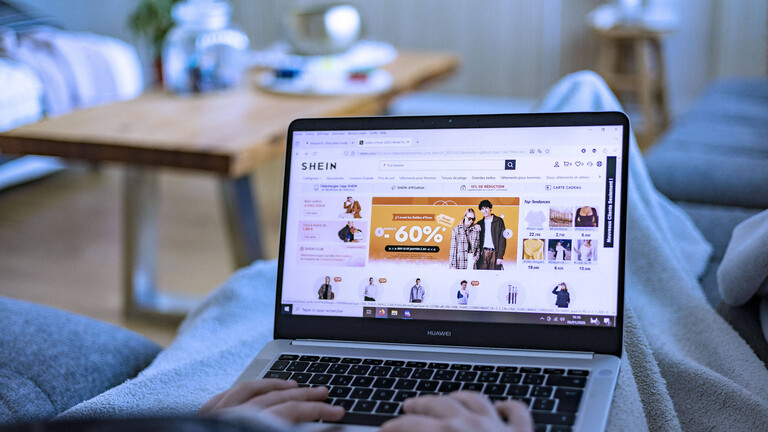 Foto von Händen, die auf einem Laptop tippen, auf dessen Bildschirm die Webseite der Shopping-Plattform Shein zu sehen ist, auf der Rabatte locken