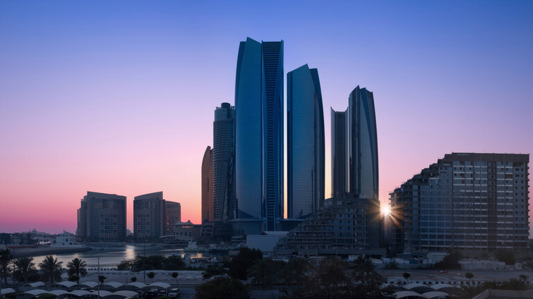Skyline von Abu Dhabi. Hinter den Hochhäusern geht die Sonne unter, der Himmel ist rosa-blau.