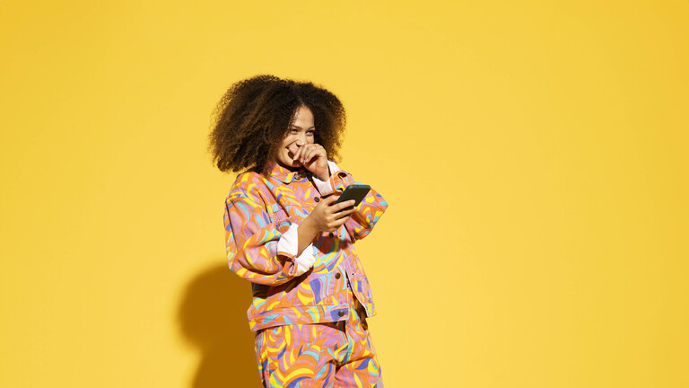 Foto einer jungen Frau un bunter Kleidung, die vor einer gelben Wand steht, ein Smartphone in der  Hand hat und sich die Hand vor den lachenden Mund hält