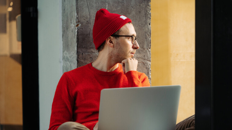 Konzentrierter freiberuflicher Designer und Autor sitzt mit seinem Laptop nachdenklich auf der Fensterbank und blickt zum Fenster hinaus.