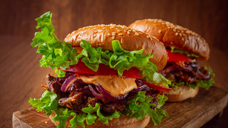 Zwei Burger mit Rindfleischstreifen, Tomaten, Röstzwiebeln und Soße, auf einem Holzbrett angerichtet; es sind keine Menschen zu sehen.