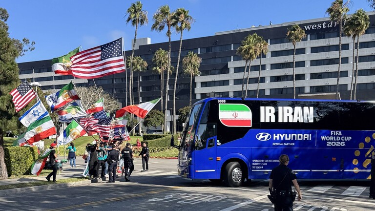 Protestierende schwenken die ehemalige iranische Flagge, die US-amerikanische und israelische Flaggen, als der Bus der Iranischen Fußballnationalmannschaft vor dem Hotel, in dem das Iranische Team untergebracht ist, wegfährt (Los Angeles, USA, 14.06.2026)