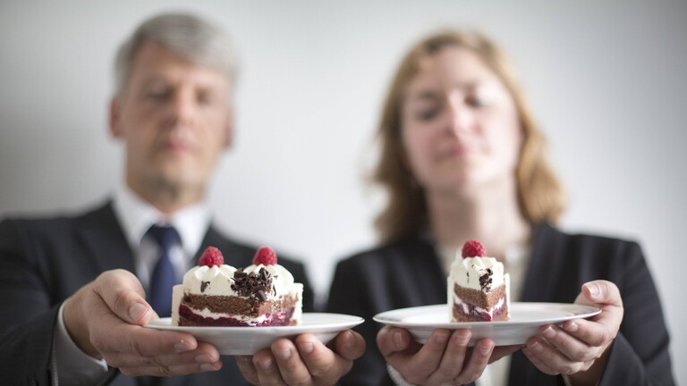Ein Mann und eine Frau stehen nebeneinander. Beide halten einen Teller in den Händen mit einem Stück Torte darauf. Das Kuchenstück auf dem Teller des Mannes ist größer als auf dem Teller der Frau.