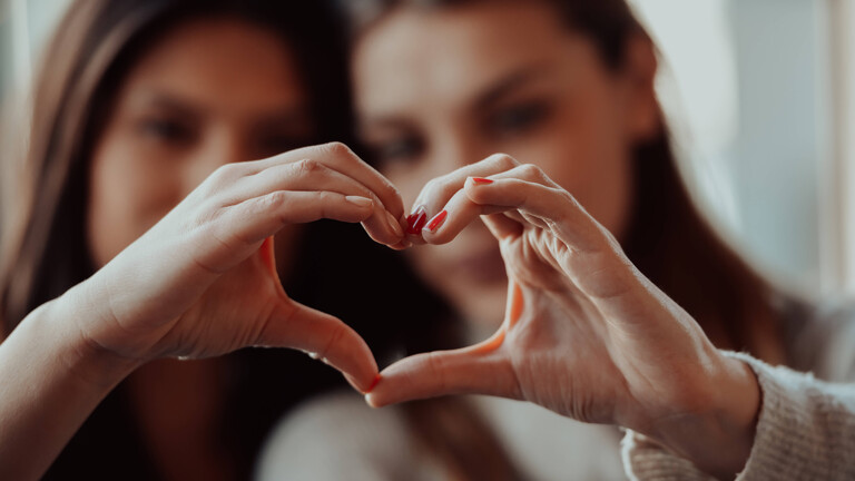 Zwei Frauen halten ihre Hände so zusammen, dass sie gemeinsam ein Herz ergeben. Die Herz-Hand ist im Vordergrund. Die beiden Frauen sind unscharf im Hintergrund.