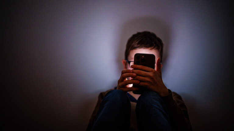 Ein Junge sitzt in einem dunklen Raum und wird von einem Smartphone beleuchtet.