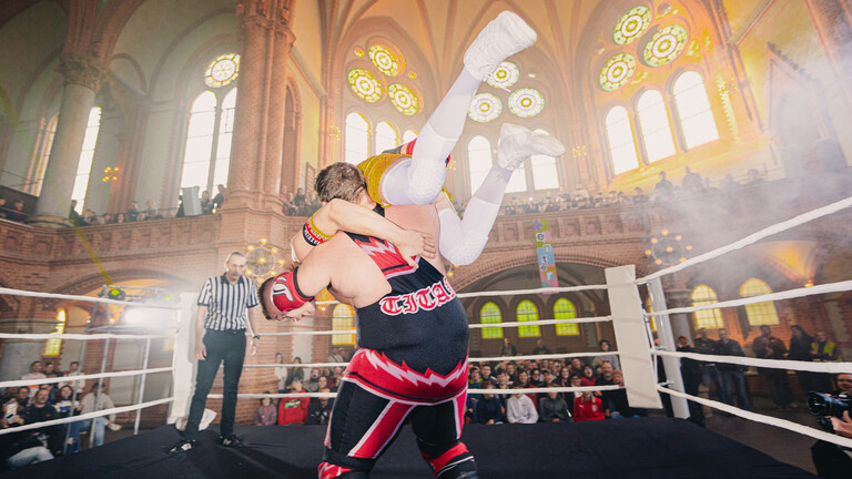 Wrestling-Veranstaltung des Vereins „enter – Junge Kulturregion Chemnitz“ // Wrestler auf Bühne mit Publikum außenherum in einer großen Halle, die wie eine ehemalige Kirche aussieht