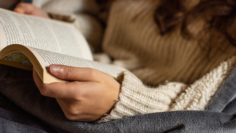 Eine junge Frau ist in eine Decke gekuschelt und liest ein Buch.