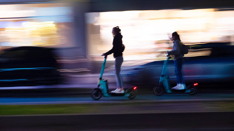 Zwei Junge Frauen fahren auf E-Scootern nach Einbruch der Dunkelheit auf einer Berliner Hauptstrasse.
