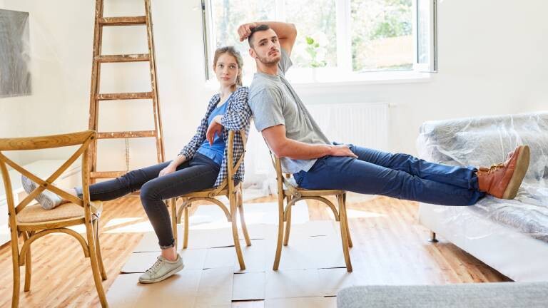 Junges Paar sitzt erschöpft und müde beim Einzug in eine neue Wohnung auf zwei Stühlen, die auf ausgefalteten Pappkartons stehen. Hinten an der Wand ist eine Leiter angelehnt. Das Sofa ist noch in Folie eingepackt.