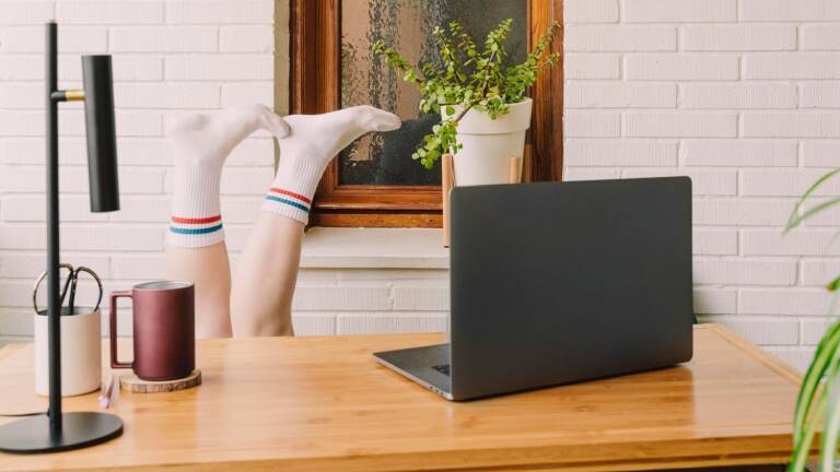 Füße mit weißen Tennissocken ragen über Schreibtisch, auf dem ein Laptop steht