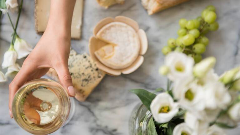Eine Frau greift über eine Tisch mit verschiedenen Käsesorten und Baguette nach einem Glas mit Weißwein.