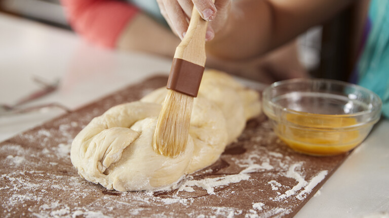 Hefeteig liegt zu einem Zopf geflochten auf einer bemehlten Arbeitsfläche. Jetzt wird der Teig mit Eigelb bestrichen.