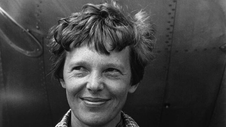 Eine Schwarz-Weiß-Nahaufnahme der lächelnden Flugpionierin Amelia Earhart mit kurzen, leicht zerzausten Haaren vor dem metallischen Rumpf ihres Flugzeugs.