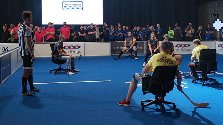 Auf einem Spielfeld in einer Halle sitzen Menschen auf Bürostühlen und mit Schlägern in der Hand. Daneben steht ein Schiedsrichter.