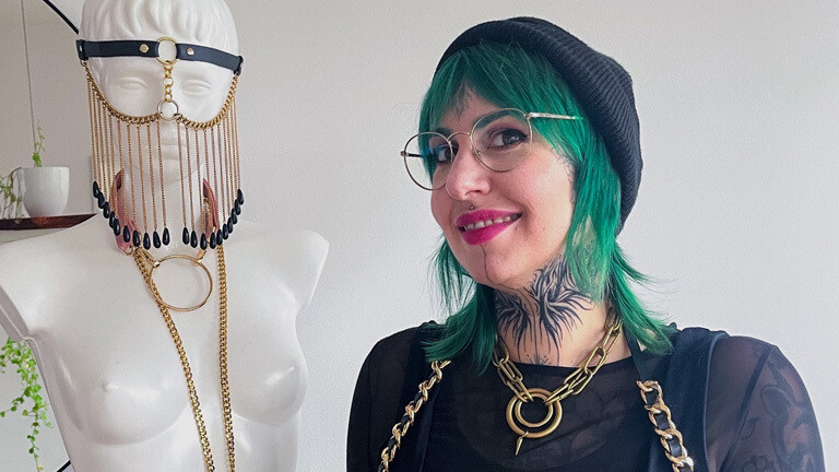 Sara Bischoff hat grüne Haare, sie trägt eine schwarze Mütze und ein schwarzes Kleid und viel Schmuck. Neben ihr ist eine Kleiderpuppe, die ebenfalls Schmuck trägt.