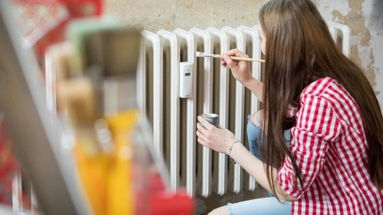 Eine junge Frau streicht in einer Wohnung eine Heizung mit weißer Farbe