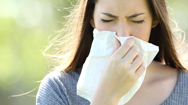 Eine Frau niest in ein Taschentusch. Sie ist draußen, im Hintergrund ist es grün.
