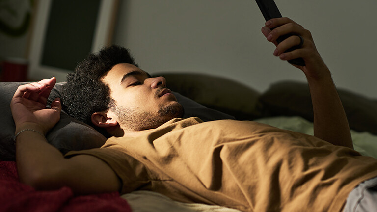 Ein Mann liegt auf dem Rücken im Bett. Sein linker Arm ist aufgestützt und er hält ein Smartphone in die Höhe, auf das er blickt. Es wirkt ruhig und etwas traurig.