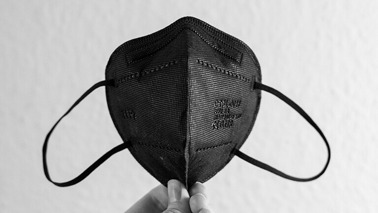 Schwarz-weiß-Foto: Die Fingerspitzen einer Hand halten eine schwarze FFP2-Maske fest.
