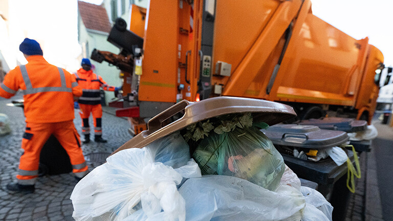 In der Stadt Reutlingen steht ein orangener Müllwagen in einer kleinen Straße, zwei Müllwerker entleeren Tonnen. Eine Bio-Tonne steht davor, darin sind befüllte Plastikbeutel, die nicht in die Tonne gehören.