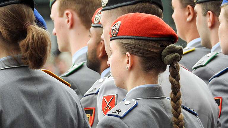 Soldaten und Soldatinnen in Uniform stehen nebeneinander, sie sind von hinten zu sehen. Eine Soldatin hat einen langen Zopf. Alle tragen Baretts. Aufnahme vom militärischen Appell bei der Parade zum Veteranentag am 15. Juni 2025.