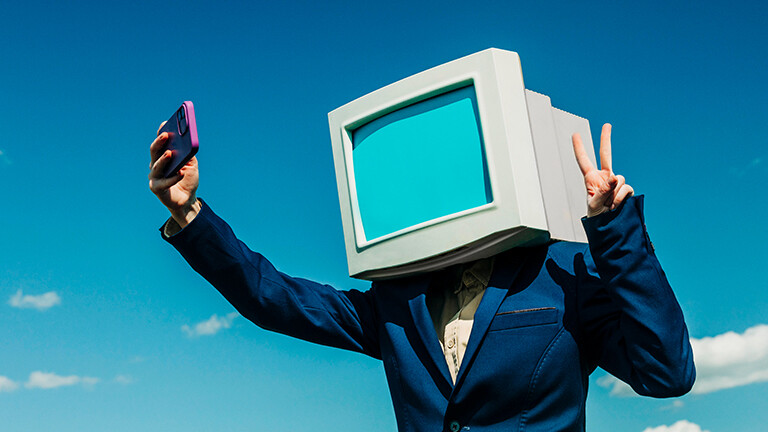 Eine Person in einem blauen Anzug trägt auf den Schultern einen Computerbildschirm, es ist eine Attrappe. Die Person macht ein Selfie von sich mit einem Smartphone.