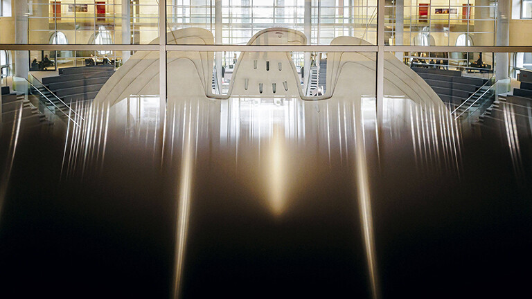 Blick durch eine Glasfront von hinten auf den Bundesadler im Plenum des Bundestages.