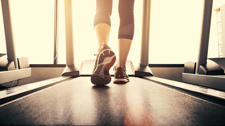 Eine Frau steht auf einem Laufband und läuft; sie trägt Turnschuhe und eine Leggings. Sie ist von hinten zu sehen. Durch das Fenster vor ihr fällt Sonnenlicht.