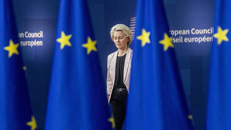 EU-Kommissionspräsidentin Ursula von der Leyen bei einer Konferenz; sie läuft hinter EU-Flaggen entlang. Die Flaggen und auch der Raum sind blau.