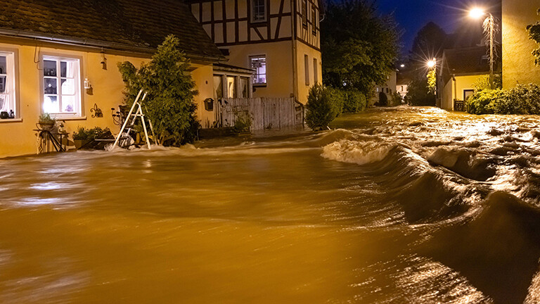 Nach starken Regenfällen steht der Ortskern des Frankfurter Stadtteils Niederursel unter Wasser. Es ist Abend, durch eine kleine Straße läuft viel Wasser, an den Seiten stehen Fachwerkhäuser.