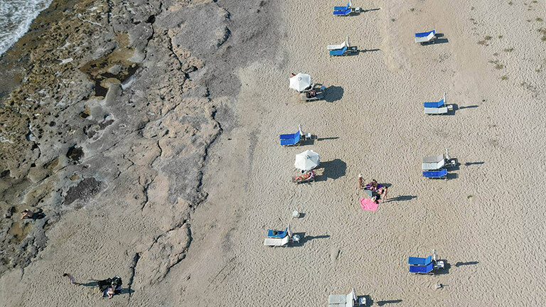 Blick aus der Vogelperspektive: Auf Zypern stehen am Strand Liegestühle mit Sonnenschirmen, es sind wenige Menschen am Strand. Es wirkt sehr heiß.