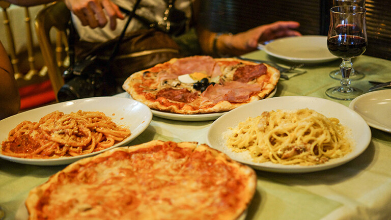 Auf einem Tisch im Restaurant stehen Teller mit Essen, mit Pizza und Nudeln. Alles wirkt sehr lecker. Eine Person sitzt am Tisch.