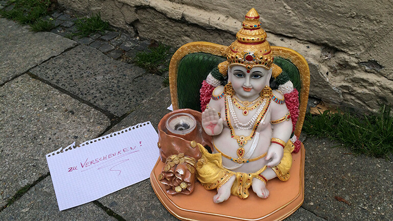 Auf einem Bürgersteig steht eine indische Heiligen-Figur in bunten Farben, daneben liegt ein Zettel auf dem handgeschrieben steht: zu verschenken.
