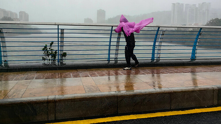 Der Taifun Kajiki zieht über China und wird auch Vietnam treffen. In der chinesischen Stadt Sanya steht eine Person auf einer Brücke und hält sich am Geländer fest. Die rosa Regenjacke flattert im Wind. Die Stadt ist in graue Wolken gehüllt. Es wirkt sehr stürmig.