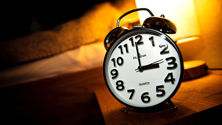 Ein Wecker steht neben einem Bett auf einem kleinen Tisch. Darauf steht auch eine Lampe, die warmes Licht macht. Der Wecker zeigt 3 Uhr.