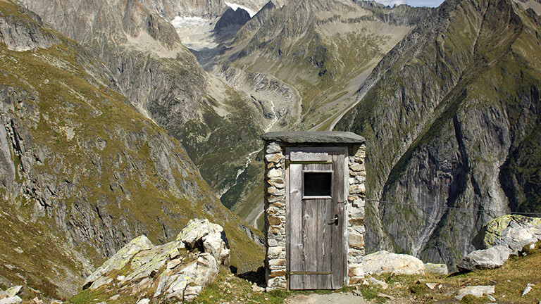 In den Schweizer Alpen steht ein kleines Toilettenhaus aus Stein und Holz.
