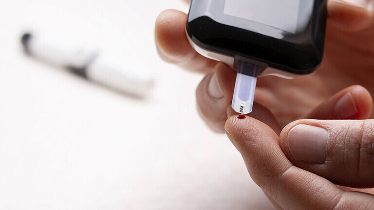 Zwei Hände: Die eine hält ein Blutzuckermessgerät an den Zeigefinger der anderen Hand. Der Finger wird leicht gepikst. Ein Blutstropfen ist auf dem Finger. Der Blutzuckerwert wird gemessen.