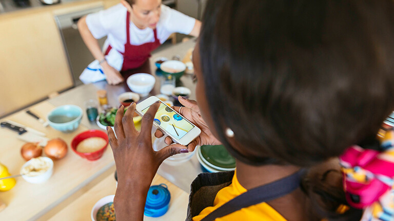 Eine Frau ist von oben zu sehen, wie sie ein Foto mit dem Smartphone von einem gedeckten Tisch macht. Auf dem Tisch stehen verschiedene Speisen in bunten Schälchen. Eine Frau sitzt am Tisch und trägt ein rote Schale.
