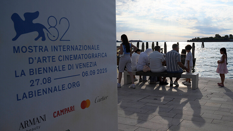 Touristen sitzen in Venedig am Wasser, die Sonne scheint. Links hängt ein Plakat, das auf die 82. Filmfestspiele in Venedig hinweist.