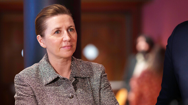 Die dänische Ministerpräsidentin Mette Fredriksen steht in einem Raum, im Hintergrund sind Personen erkennbar. Sie blickt nach rechts. Sie trägt eine braune Stoffjacke.