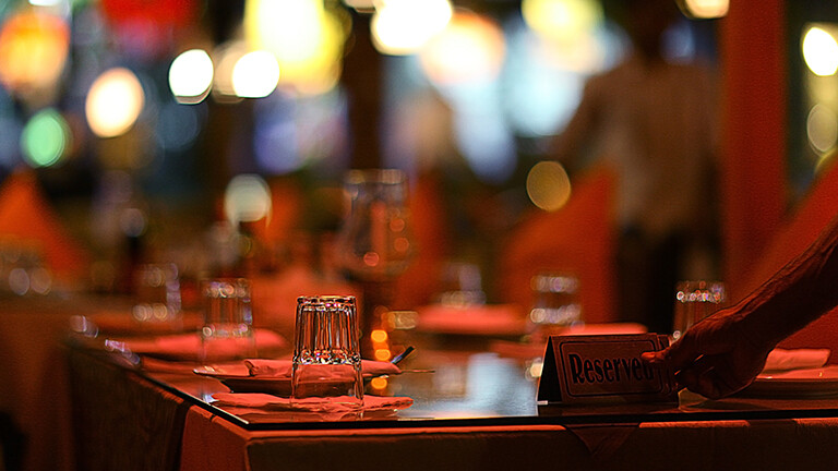 In einem Restaurant steht ein gedeckter Tisch. Eine Hand ist zu sehen, die ein Schild auf den Tisch stellt mit "reserved". Es herrscht sehr angenehmes Licht, es wirkt edel und gemütlich.