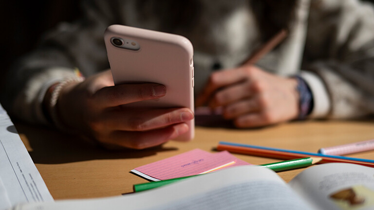 Ein Schulkind sitzt am Schreibtisch, bearbeitet Aufgaben mit einem Stift und hält dabei ein Smartphone in der Hand.