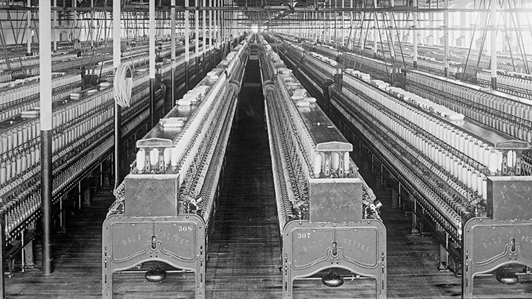Schwarz-weiß-Foto: Blick in eine Baumwollspinnerei zwischen 1909 und 1920 in Manchester; eine große, menschenleere Fabrikhalle voller Webstühle.