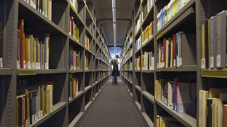 Blick in eine Bibliothek. Rechts und links stehen lange Regalreihen mit Büchern. Am Ende des Ganges steht eine Person.