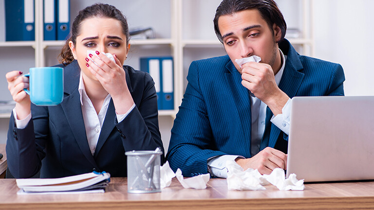 Symbolfoto: Ein Mann und eine Frau sitzen hinter einem Schreibtisch. Sie trägt einen blauen Blazer, er eine blaue Anzugsjacke. Sie halten sich Taschentücher vor den Mund und wirken sehr krank. Die Situation wirkt grotesk-lustig.