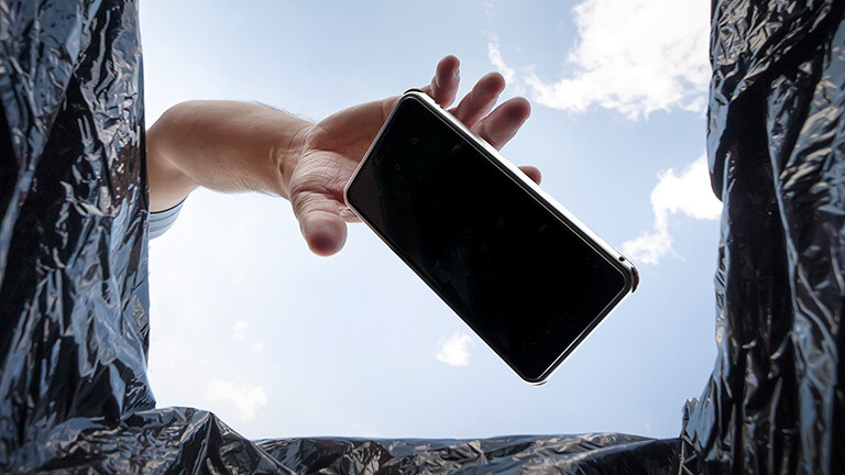 Blick aus einem Mülleimer in den blauen Himmel. Eine Hand wirft ein Smartphone in den Müll.