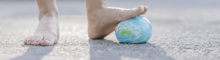 Zwei nackte Füße; der linkte drückt einen Erdkugel aus Stoff nach unten. Von hinten fällt Licht, sodass die Füße einen Schatten werfen.